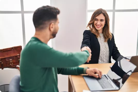 Mulher sorridente e homem apertando as mãos em um ambiente de escritório com notebooks abertos sobre a mesa, simbolizando fechamento de contrato ou início de parceria.