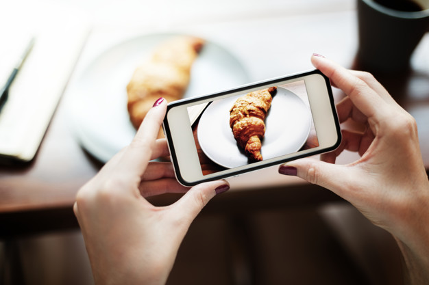 duas mãos segurando um celular e tirando foto de um croissant