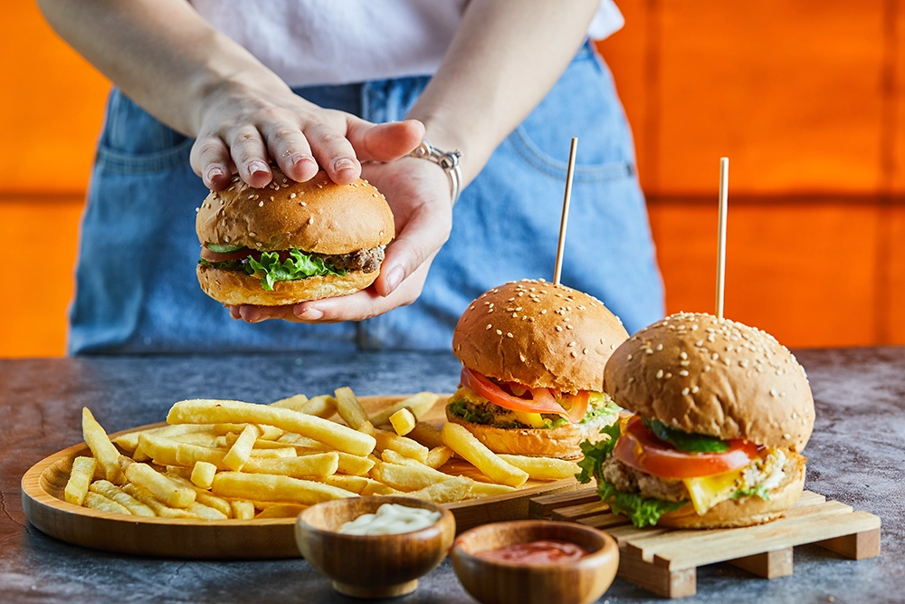 mulher segurando um hamburguer em cima de uma mesa com hamburgueres e batatinhas