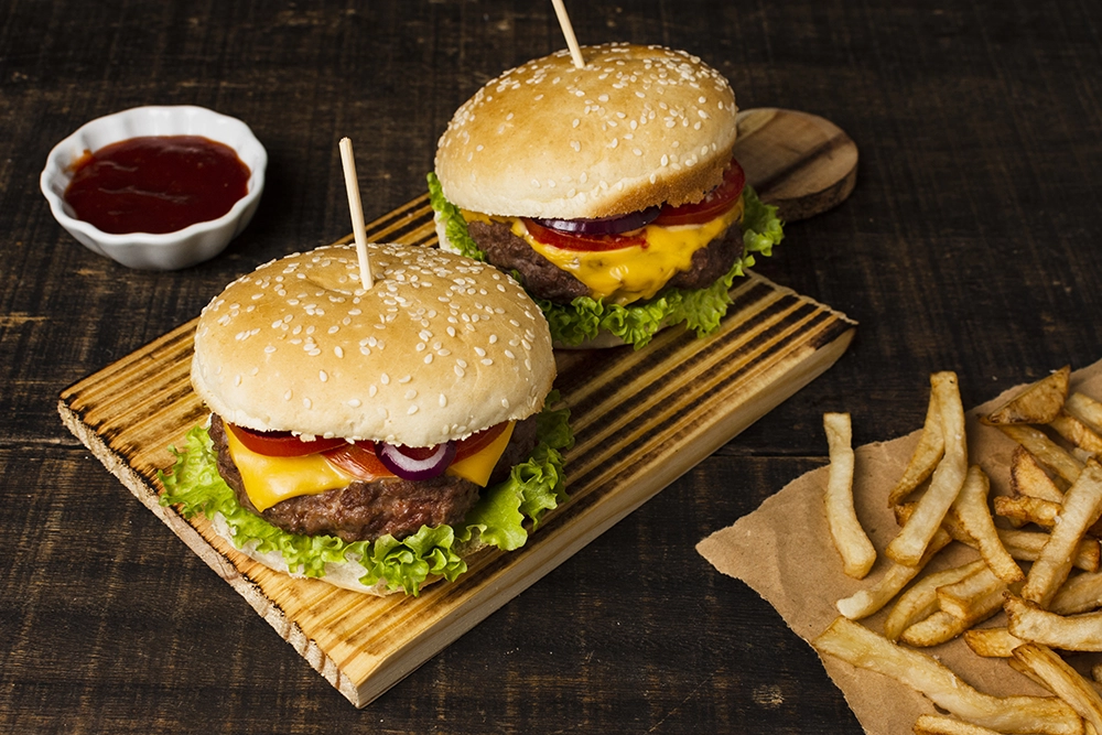 Dois hamburgueres apoiado em uma tabua de madeira em cima de uma mesa acompanhado de batatas fritas