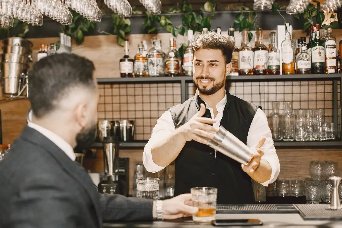 Barman sorridente preparando um drink enquanto interage com cliente em um bar elegante.