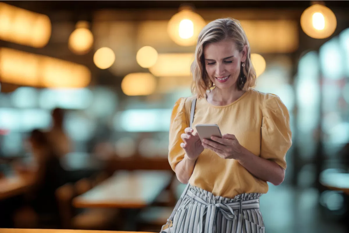 Mulher loira sorrindo enquanto usa o smartphone em pé no salão de um restaurante com iluminação quente e mesas ao fundo