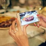 Mãos femininas fotografando com smartphone um prato de carne com molho e acompanhamentos sobre mesa posta em restaurante, com a foto aparecendo na tela do celular