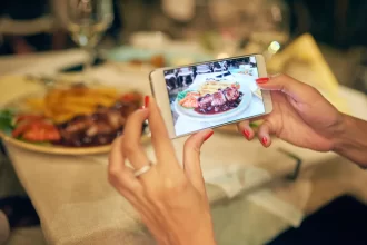 Mãos femininas fotografando com smartphone um prato de carne com molho e acompanhamentos sobre mesa posta em restaurante, com a foto aparecendo na tela do celular
