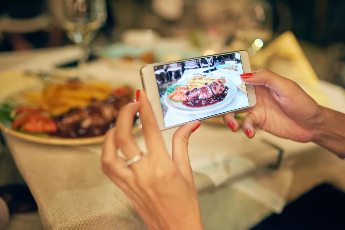 Mãos femininas fotografando com smartphone um prato de carne com molho e acompanhamentos sobre mesa posta em restaurante, com a foto aparecendo na tela do celular