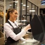 Funcionária de padaria com avental escuro e luvas descartáveis operando o sistema PDV touchscreen no balcão de atendimento, com prateleiras de pães ao fundo, representando o registro de sangria de caixa no sistema durante o expediente
