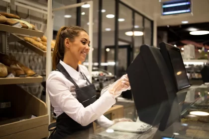 Funcionária de padaria com avental escuro e luvas descartáveis operando o sistema PDV touchscreen no balcão de atendimento, com prateleiras de pães ao fundo, representando o registro de sangria de caixa no sistema durante o expediente