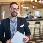 Gestor de restaurante de terno e camisa xadrez segurando caderno espiral, de pé em frente ao balcão do bar com banquetas e funcionários ao fundo