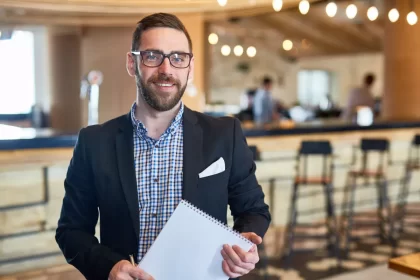Gestor de restaurante de terno e camisa xadrez segurando caderno espiral, de pé em frente ao balcão do bar com banquetas e funcionários ao fundo