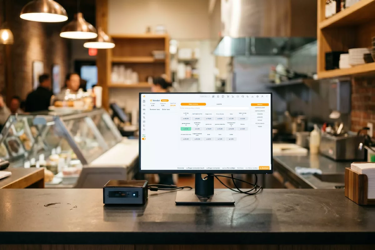 mini PC compacto ao lado de monitor no balcão de um bar com o sistema ControleNaMão aberto na tela, ambiente de restaurante desfocado ao fundo