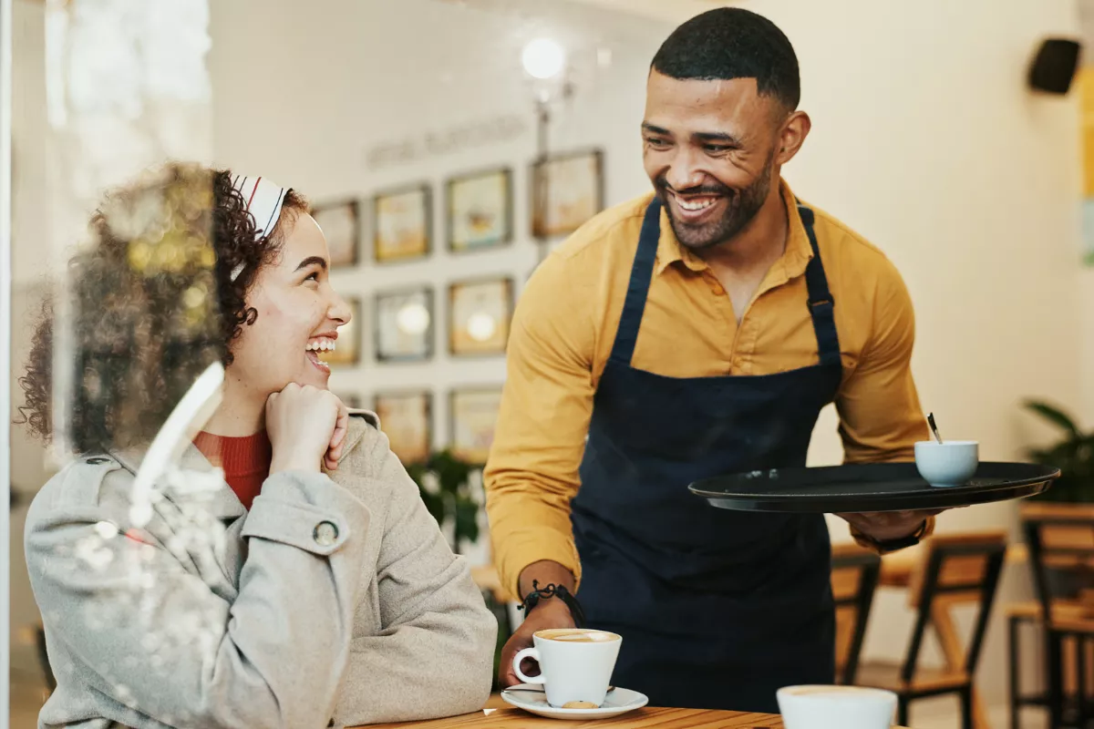 Garçom com avental escuro segurando bandeja com cafés enquanto sorri para cliente sentada à mesa de cafeteria iluminada, em cena de atendimento descontraído