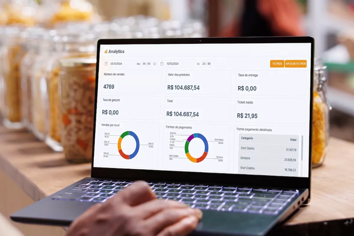 Mãos digitando em notebook com dashboard do CNM Analytics aberto, exibindo número de vendas, ticket médio, faturamento total e gráficos de formas de pagamento, com potes de ingredientes ao fundo