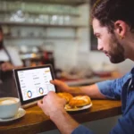 Homem barbudo de camisa azul consultando dashboard do CNM Analytics em tablet no balcão de uma cafeteria, com barista de avental ao fundo e xícara de café e pão na bancada