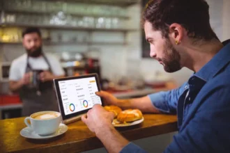 Homem barbudo de camisa azul consultando dashboard do CNM Analytics em tablet no balcão de uma cafeteria, com barista de avental ao fundo e xícara de café e pão na bancada