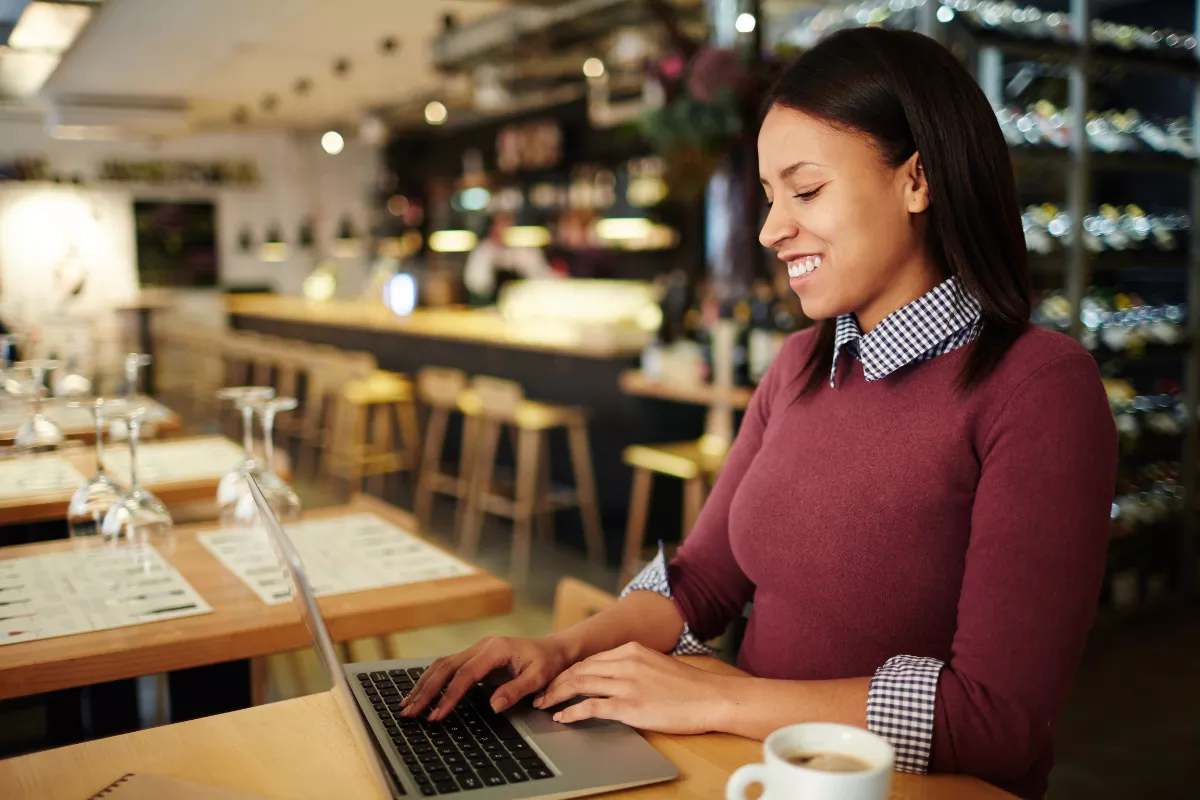 Gestora de restaurante de blusa vinho sorrindo ao usar notebook em mesa do salão vazio, com taças, cardápios e prateleiras de bebidas ao fundo