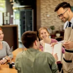 Garçom com óculos e avental bege anotando pedido e conversando com mesa de três clientes sorridentes em restaurante iluminado, com cardápio aberto na mão