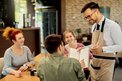 Garçom com óculos e avental bege anotando pedido e conversando com mesa de três clientes sorridentes em restaurante iluminado, com cardápio aberto na mão