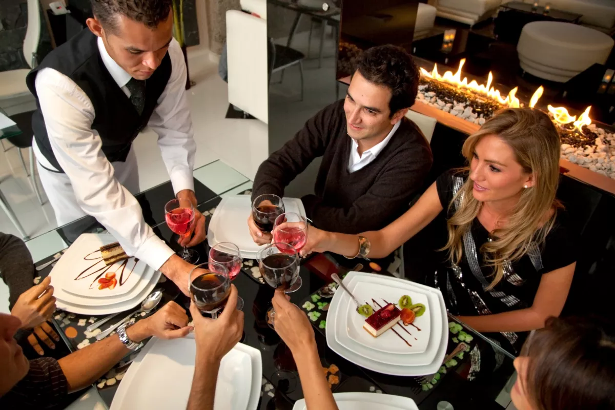 Grupo de clientes brindando com taças de vinho em restaurante sofisticado, garçom de colete participando do brinde ao lado da mesa com pratos de sobremesa