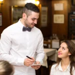 Garçom jovem de camisa branca e gravata borboleta sorrindo enquanto anota pedido de cliente feminina sentada em restaurante de madeira com mesas ao fundo