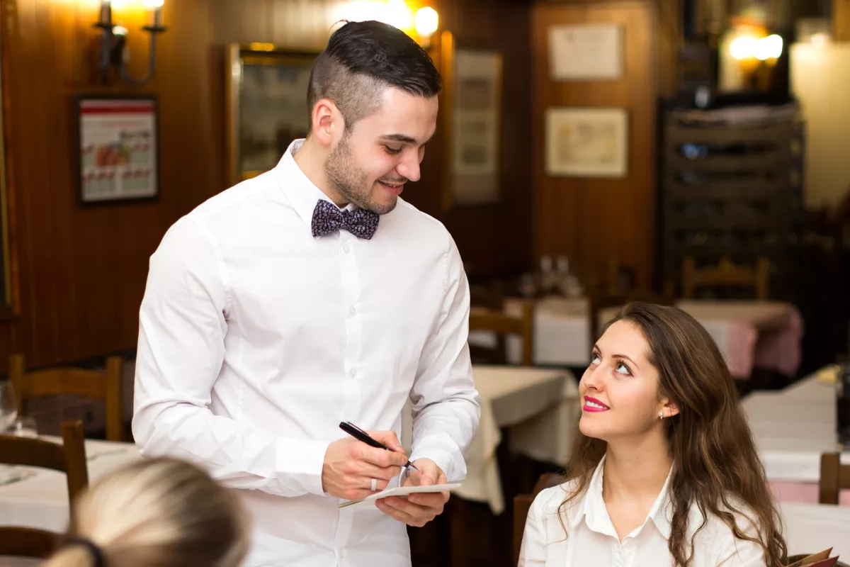 Garçom jovem de camisa branca e gravata borboleta sorrindo enquanto anota pedido de cliente feminina sentada em restaurante de madeira com mesas ao fundo