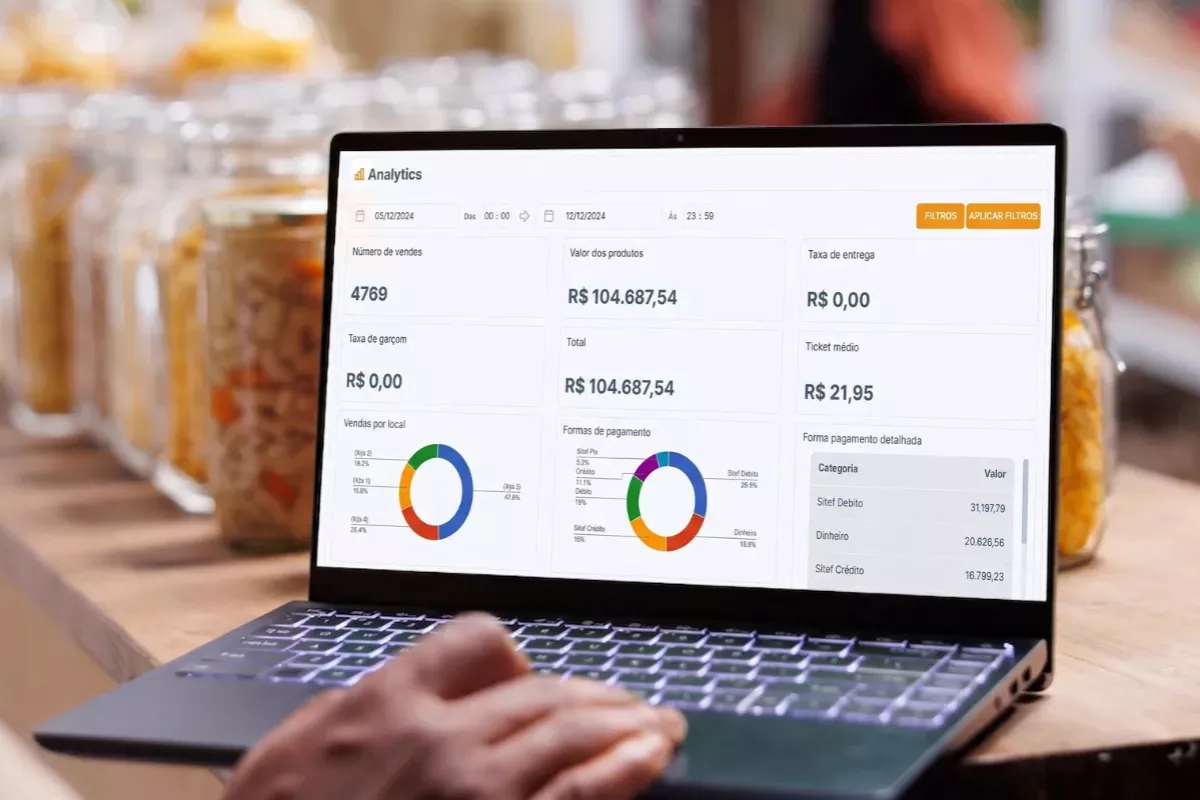 Mãos digitando em notebook com dashboard do CNM Analytics aberto, exibindo número de vendas, ticket médio, faturamento total e gráficos de formas de pagamento, com potes de ingredientes ao fundo