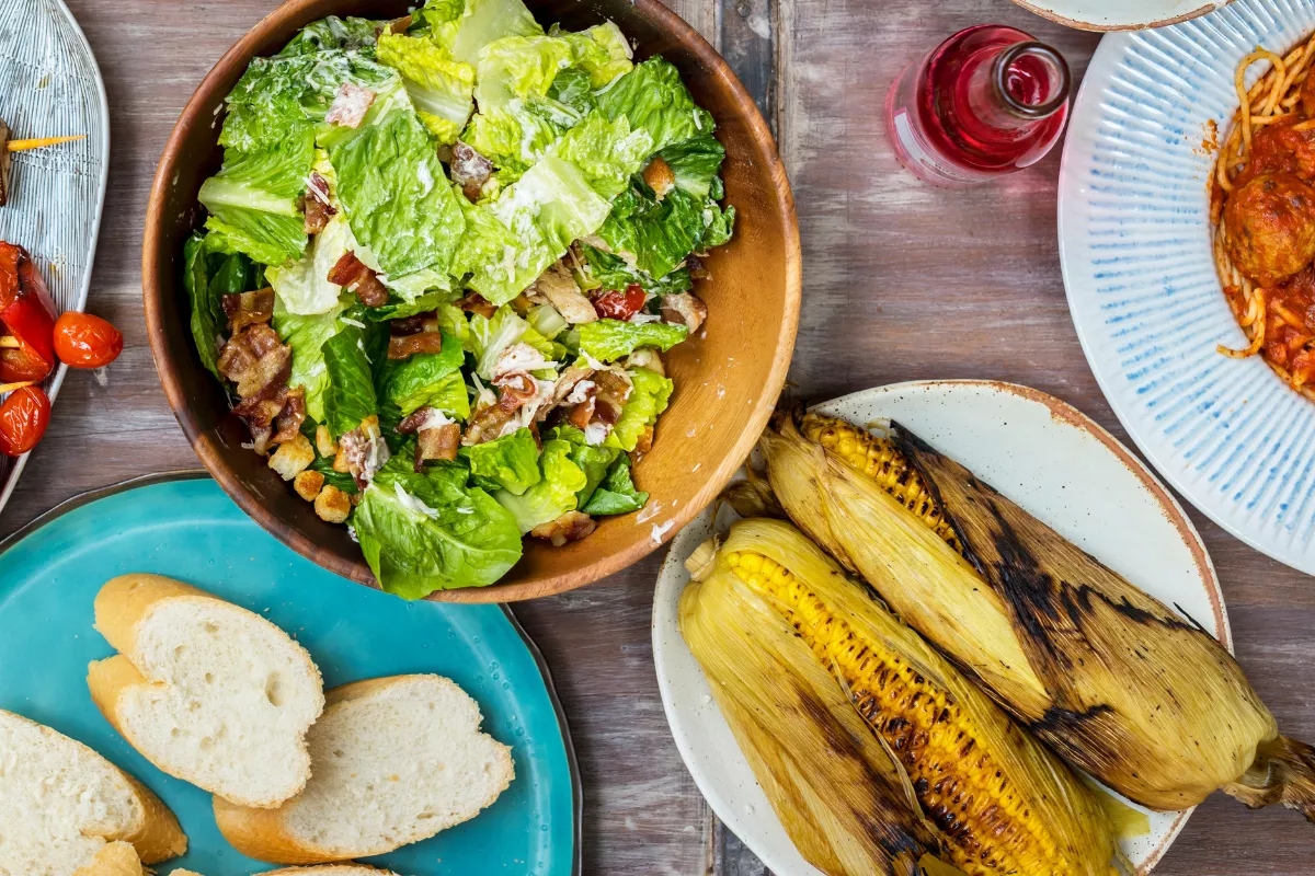 Mesa de restaurante vista de cima com salada caesar em tigela de barro, milho grelhado, pão fatiado e macarrão ao molho, ilustrando a variedade de insumos que dependem de bons fornecedores