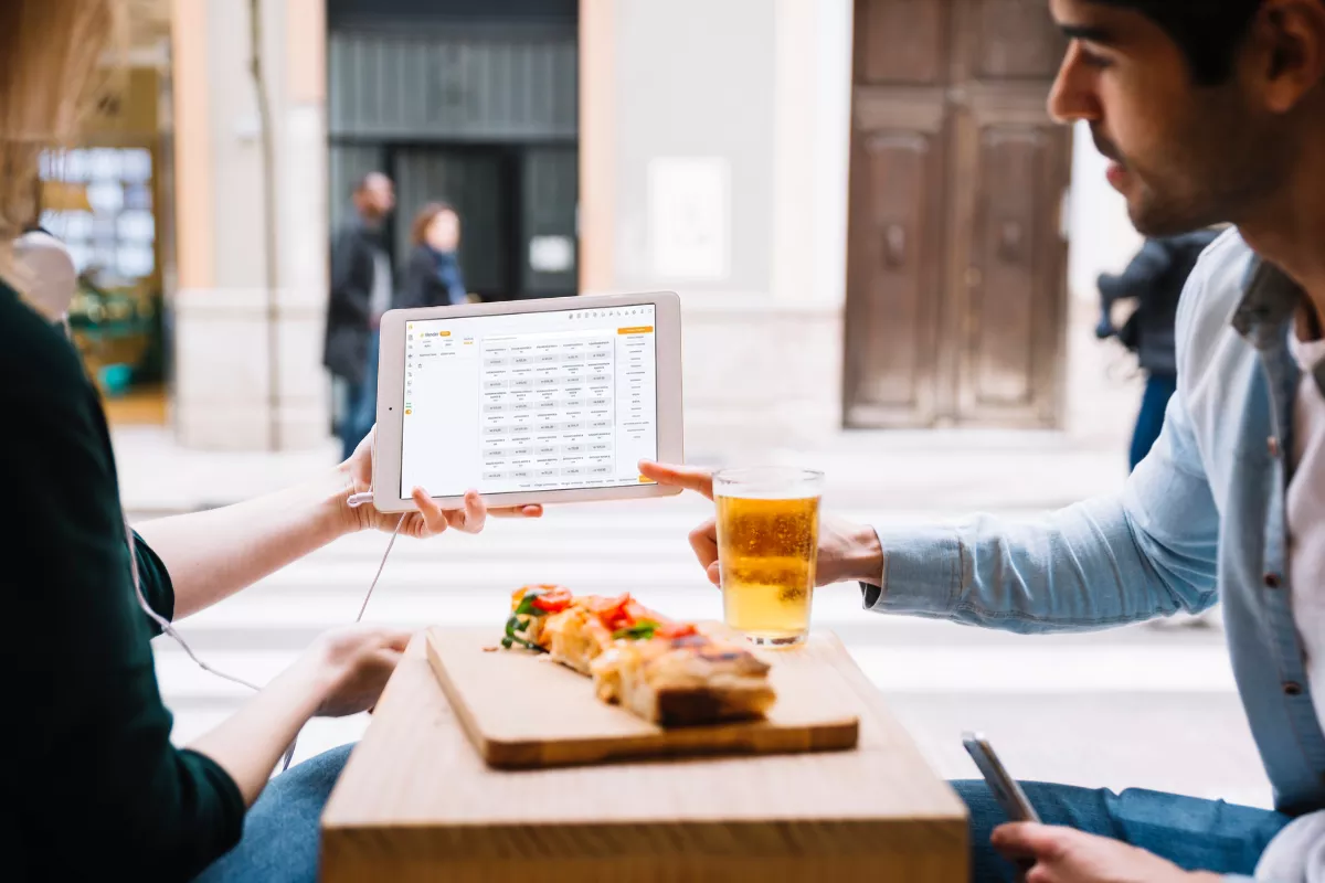 Dois clientes sentados em mesa de bar com petisco e cerveja, mostrando um ao outro a tela de um tablet com o sistema de gestão do restaurante aberto