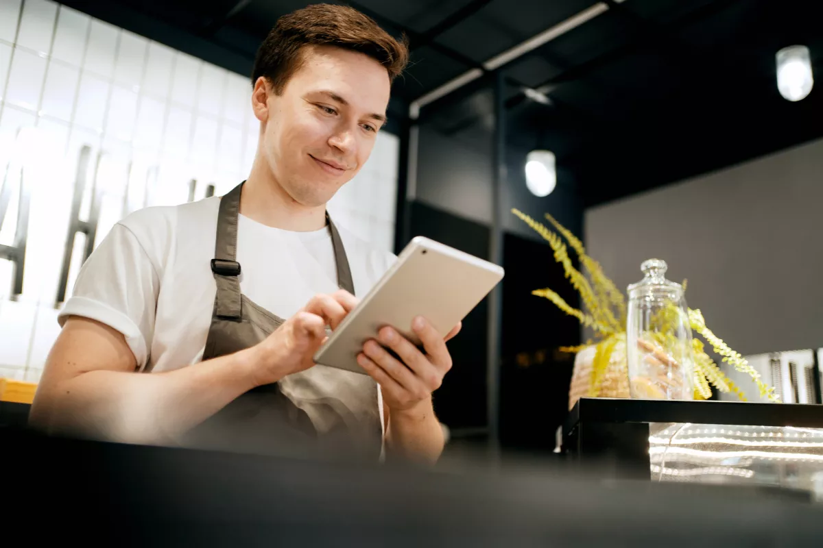 Funcionário de restaurante com avental consultando tablet no balcão do estabelecimento com expressão tranquila
