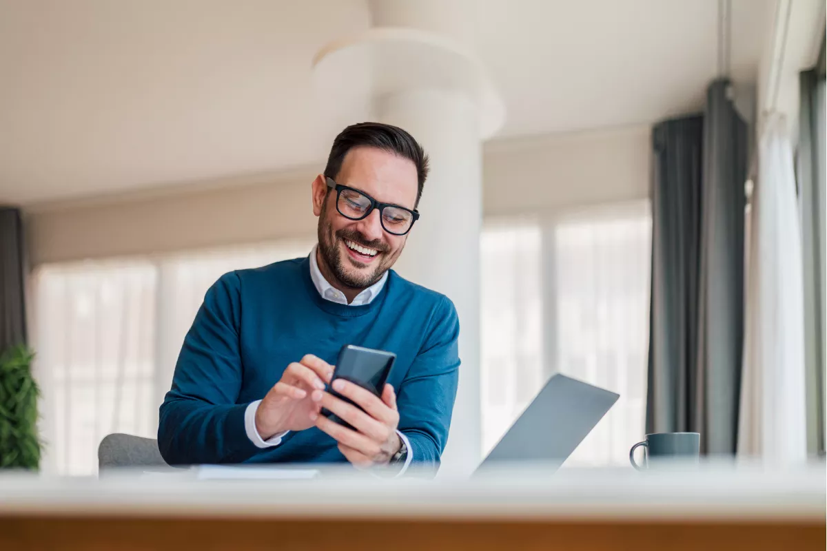 Homem de óculos e suéter azul sorrindo enquanto usa o celular sentado em casa, com notebook aberto à frente