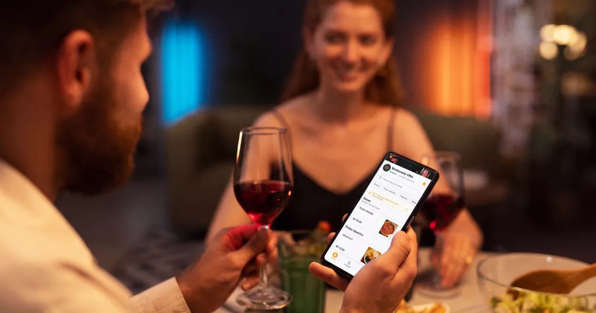 Casal em jantar romântico num restaurante iluminado, com o homem segurando celular exibindo o cardápio digital Vina enquanto brindam com taças de vinho tinto