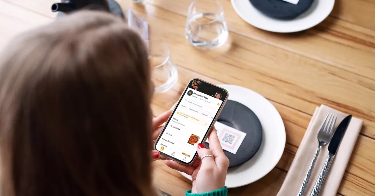 Cliente sentada à mesa de restaurante navegando no cardápio digital Vina pelo celular, com QR Code visível na mesa ao lado do prato e talheres