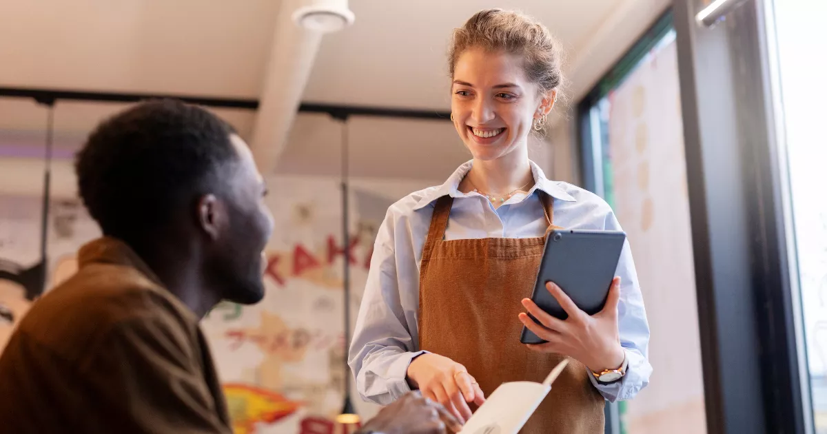 Garçonete jovem com avental marrom e tablet na mão sorri para cliente sentado, anotando pedido em caderneta, no salão iluminado de restaurante com cartazes coloridos ao fundo