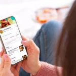 Cliente deitada no sofá em casa acessando cardápio digital de hamburgueria pelo celular, com categorias, fotos dos pratos e preços visíveis na tela