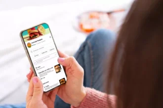 Cliente deitada no sofá em casa acessando cardápio digital de hamburgueria pelo celular, com categorias, fotos dos pratos e preços visíveis na tela