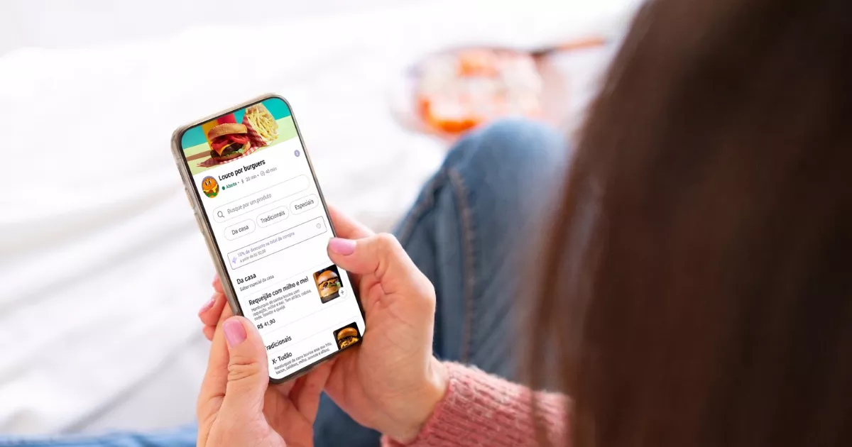Cliente deitada no sofá em casa acessando cardápio digital de hamburgueria pelo celular, com categorias, fotos dos pratos e preços visíveis na tela