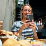 Mulher de cabelo loiro e camiseta listrada fotografando mesa de restaurante com o celular, com pratos e pães à sua frente e outro cliente ao fundo