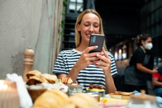 Mulher de cabelo loiro e camiseta listrada fotografando mesa de restaurante com o celular, com pratos e pães à sua frente e outro cliente ao fundo