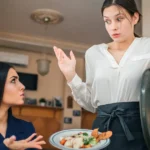 Cliente insatisfeita sentada à mesa questiona garçonete que segura prato e tampa com expressão de surpresa, no salão de restaurante com mesas e quadros ao fundo