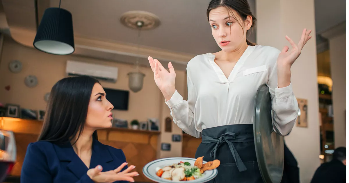 Cliente insatisfeita sentada à mesa questiona garçonete que segura prato e tampa com expressão de surpresa, no salão de restaurante com mesas e quadros ao fundo