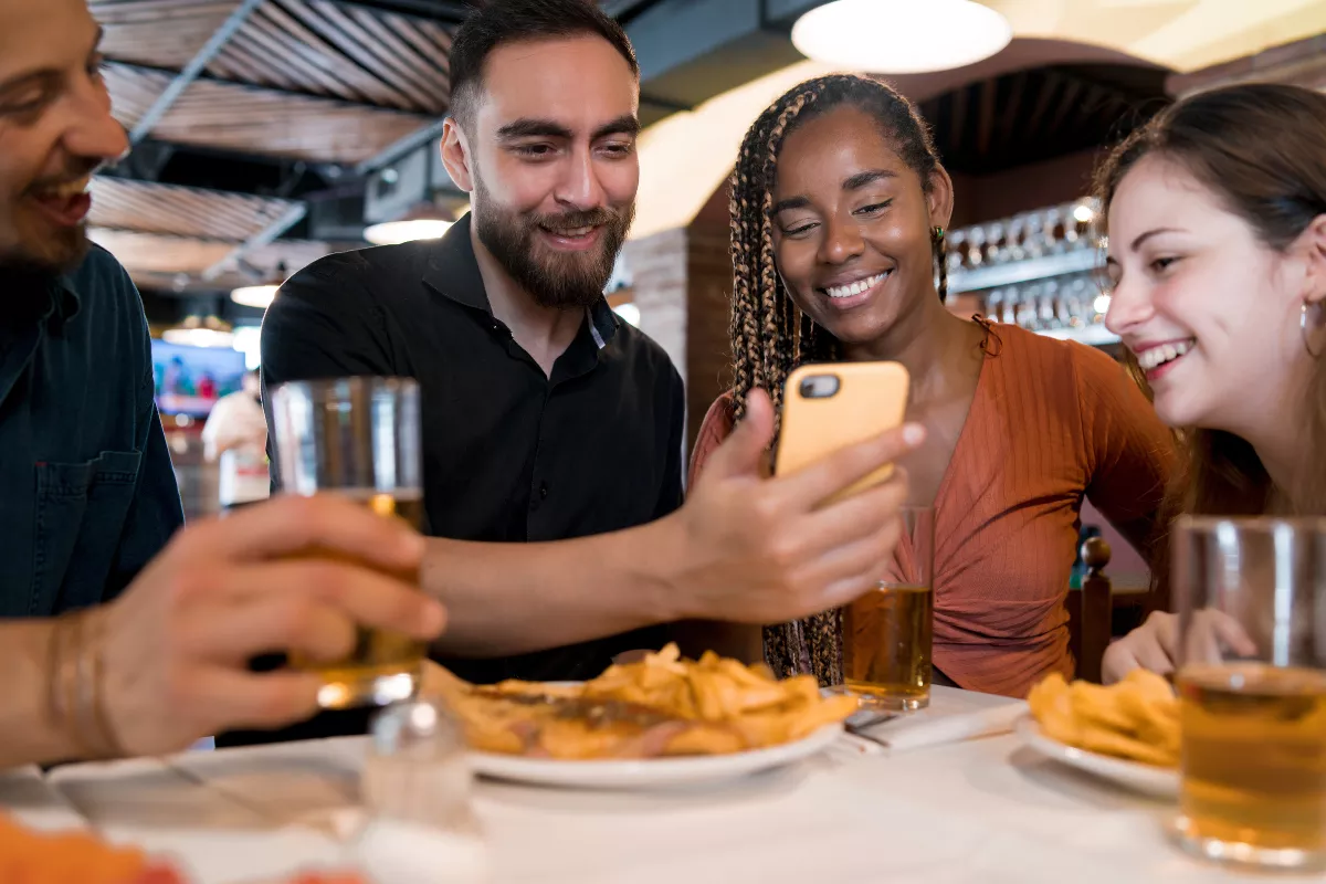 Grupo de quatro amigos sentados em bar, sorrindo enquanto olham juntos para a tela do celular, com cervejas e batatas fritas na mesa