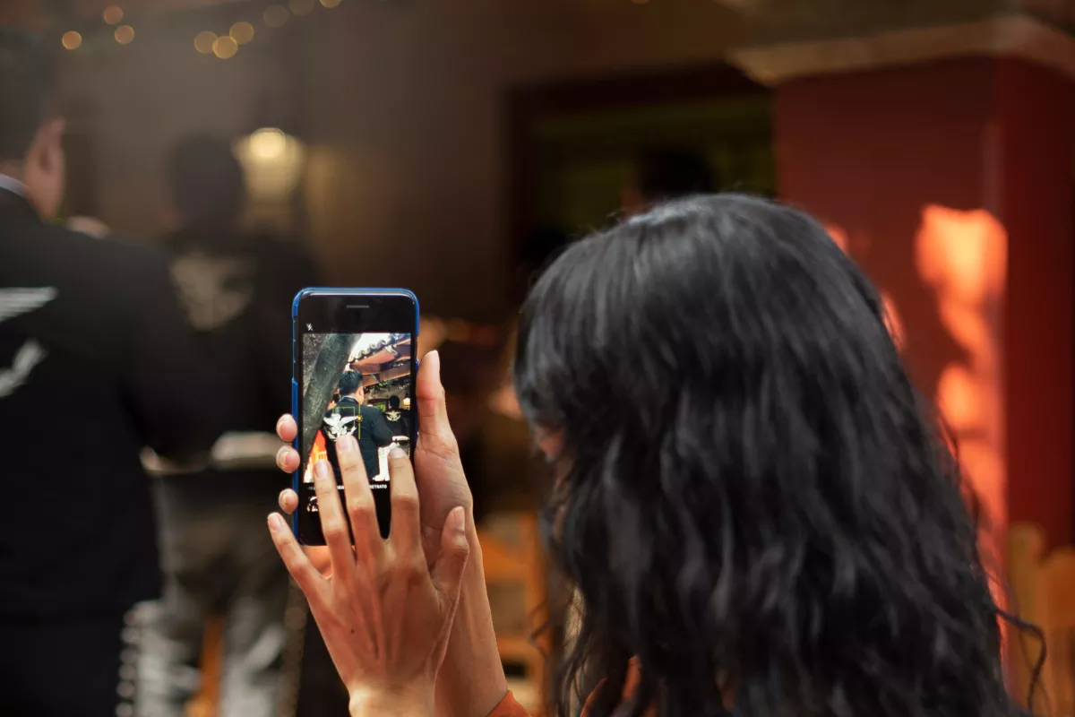 
Cliente de cabelo escuro filmando o salão do restaurante pelo celular, com luzes de decoração e outro cliente ao fundo em ambiente de iluminação quente