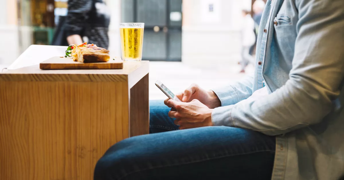 Cliente sentado mexendo no celular ao lado de uma tábua de madeira com lanche e copo de cerveja, em área aberta de restaurante com movimento ao fundo