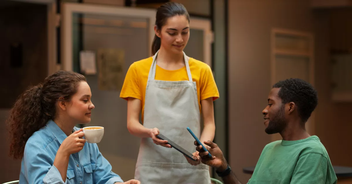 Garçonete de avental cinza e camiseta amarela mostrando tablet para casal sentado à mesa, com cliente segurando xícara de café em restaurante com fundo desfocado