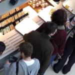 Vista aérea de grupo de clientes aguardando atendimento em balcão de lanchonete com sanduíches, frutas e sucos expostos nas prateleiras iluminadas