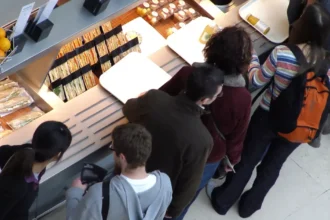 Vista aérea de grupo de clientes aguardando atendimento em balcão de lanchonete com sanduíches, frutas e sucos expostos nas prateleiras iluminadas