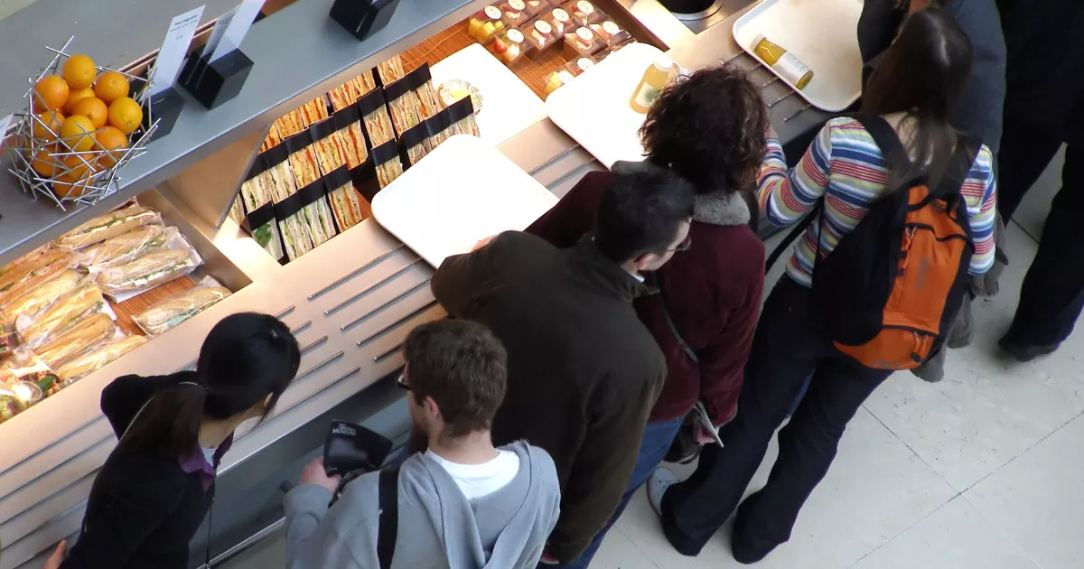 Vista aérea de grupo de clientes aguardando atendimento em balcão de lanchonete com sanduíches, frutas e sucos expostos nas prateleiras iluminadas
