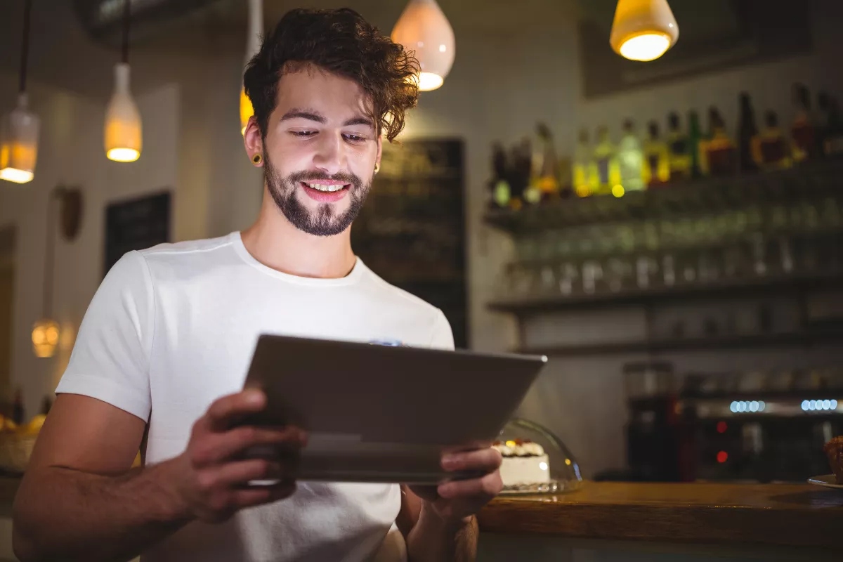 Jovem sorrindo enquanto usa tablet no balcão de um bar, com prateleiras de bebidas e iluminação quente ao fundo, representando o uso de sistema de gestão no dia a dia do restaurante