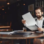 Dono de bar sentado em mesa redonda de madeira revisando documentos com caneta na mão, caderno e bebida sobre a mesa, com o salão escuro ao fundo, representando o planejamento da abertura do restaurante
