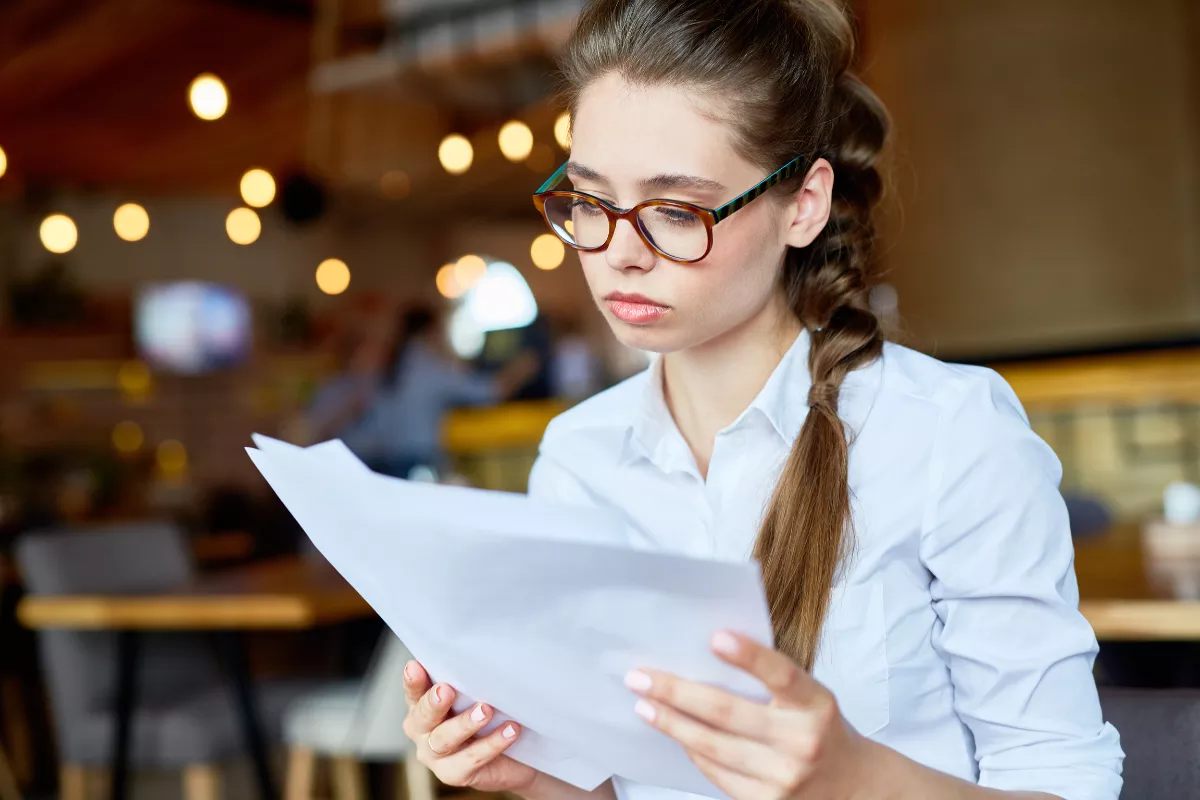Jovem com óculos e trança lendo documentos com expressão concentrada dentro de um restaurante com iluminação quente ao fundo, representando a análise da documentação para abertura de estabelecimento