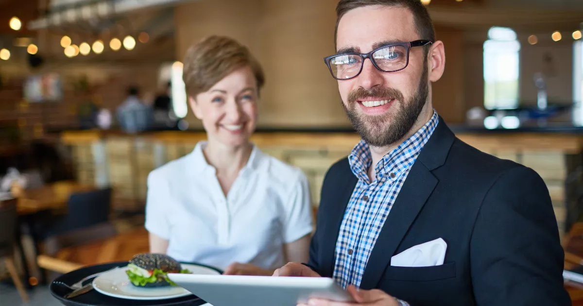 Gestor de restaurante com óculos e blazer segura tablet e sorri para a câmera ao lado de garçonete com bandeja, no salão iluminado do estabelecimento